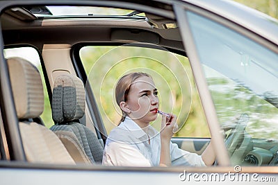 Concept of danger driving. Young woman driver red haired teenage girl painting her lips doing applying make up while driving the Stock Photo