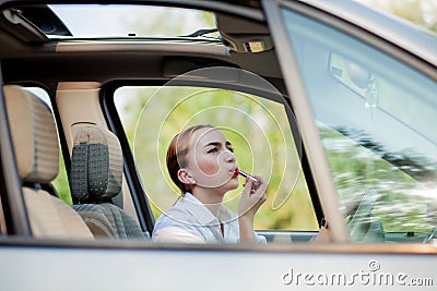Concept of danger driving. Young woman driver red haired teenage girl painting her lips doing applying make up while driving the Stock Photo