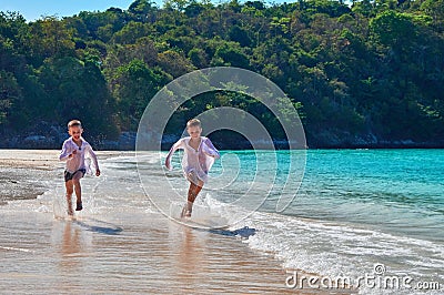 Concept entertainment children on the beach. Two laughing boys run on the turquoise sea beach on background the tropical jungle. Stock Photo