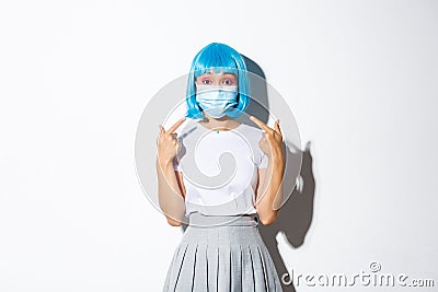 Concept of halloween celebration and covid-19 pandemic. Image of asian girl in blue wig pointing at medical mask Stock Photo