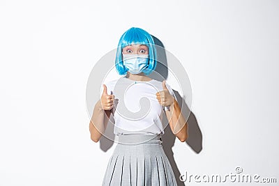 Concept of halloween during covid-19 pandemic. Image of surprised asian woman in blue wig and medical mask showing Stock Photo