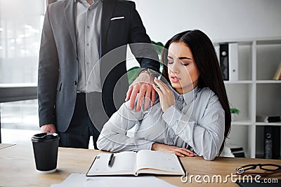 Concept harassment. Afraid young woman sit at table. Harrasment goes out of her boss. He keep hand on her shoulder. Guy Stock Photo