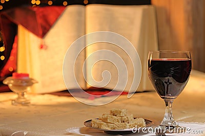 Concept Lord`s Supper and Christian communion. Bread and wine in chalice are front of big Bible. Cross reflect in goblet, candle Stock Photo