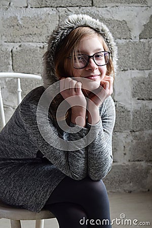 Concept portrait of a pleasant friendly happy teenager in glasses on chair. Young girl is sitting in a gray dress and smiling Stock Photo