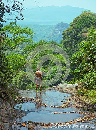 Man in swimming trunks stands at the edge of waterfall Stock Photo