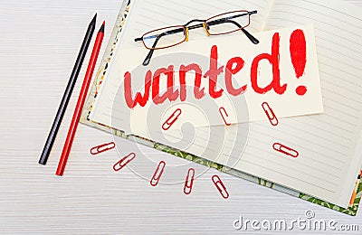 Conceptual hand drawn inscription:Wanted on the signboard.Red painting stroke sketch.Open notebook with pencils and clips,glasses. Stock Photo