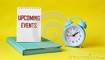 Conceptual message UPCOMING EVENTS in notepad and on yellow background, next to alarm clock and green diary. Stock Photo
