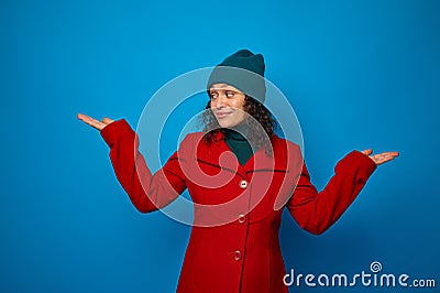 Conceptual portrait of a charming smiling curly haired woman in bright red coat and woolen green hat poses against blue background Stock Photo