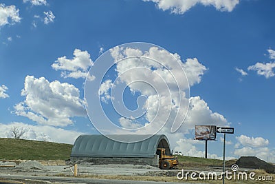 Concrete and tent housing for road maintaniance equipment with yellow tractor and piles of dirt and sand near highway in USA with Editorial Stock Photo