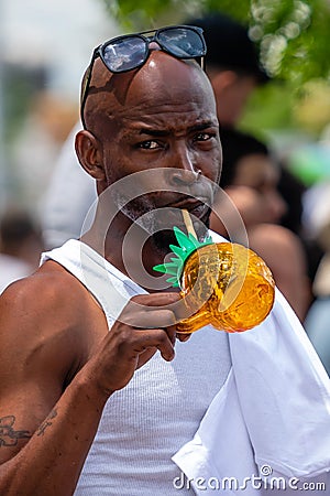 Coney Islands annual mermaid parade, an african man stands in a crowd and drinks a refreshing drink, Brooklyn, New York City, USA Editorial Stock Photo