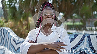 Confident african american woman joyfully stands in city park, arms crossed, sporting a beautiful, relaxed smile Stock Photo