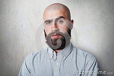 A confident bald male with thick black eyebrows and beard wearing checked shirt having gloomy expression posing against white back Stock Photo