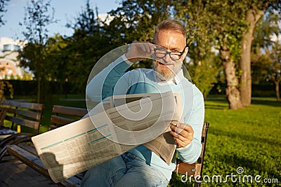 Confident bearded mature man reading daily newspaper on park bench Stock Photo