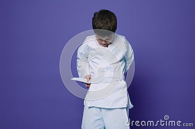 Confident Caucasian school age boy puts on white kimono, ties a belt around his waist, ready for combat sports. Aikido fighter. Stock Photo