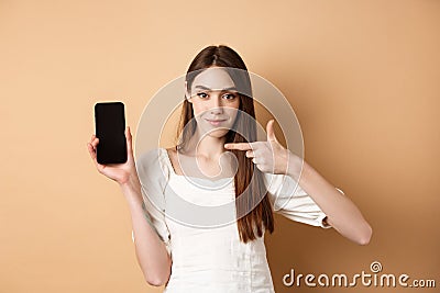 Confident girl pointing at empty smartphone screen, showing online promo deal, standing on beige background Stock Photo