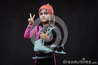 Confident little skater girl dressed in sportswear holds ice skates and shows victory hand. Isolated on a dark textured Stock Photo