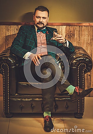 Confident old-fashioned man sitting in comfortable leather chair with glass of whisky in wooden interior at Barber shop. Stock Photo