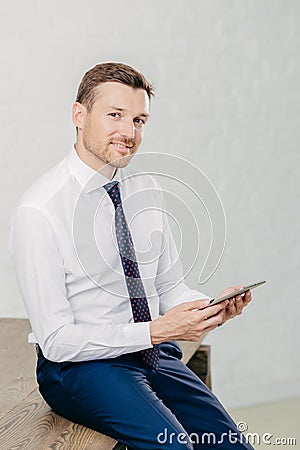 Confident pleased young male entrepreneur in luxury formal clothes, reads recieved text message from colleague on modern tablet co Stock Photo