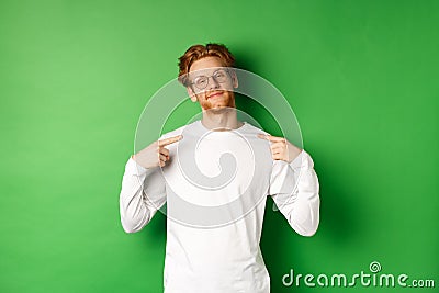 Confident and smug redhead man in glasses smiling, pointing at himself self-assured, standing over green background Stock Photo