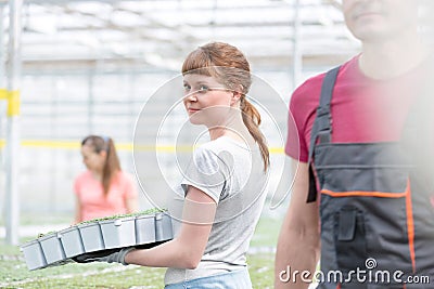 Confident young female botanist carrying seedlings by coworker in greenhouse Stock Photo