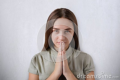 A confident young girl with beautiful dark eyes and hair praying with her hands together on grey background having believe in bett Stock Photo