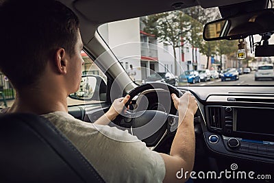 Confident young man driving car on the city streets. Teen driver keeps hands on the steering wheel looking ahead at the urban Stock Photo