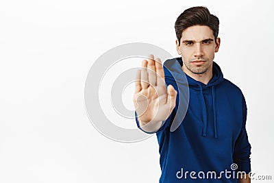 Confident young man extends hand to say stop, warning gesture, prohibit action, telling no, standing over white Stock Photo
