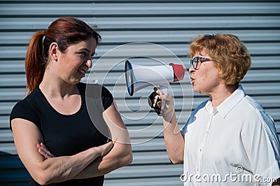 The conflict of generations. An old woman yells into a megaphone at her middle-aged daughter against gray background Stock Photo