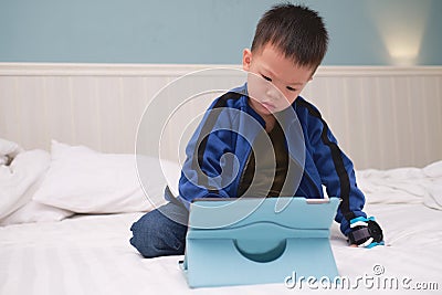 Confused aggressive Asian toddler boy child sitting in bed watching a video , playing game from tablet pc, Gadget-addicted Stock Photo