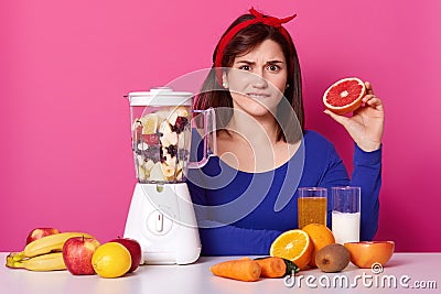 Confused black haired emotional female sits at table, biting her lip, holding half of grapefruit in one hand, mixing fruit in Stock Photo