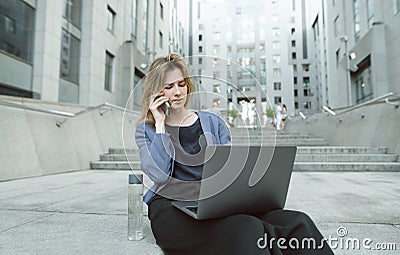 Confused businesswoman talking via smartphone looking at her laptop on the stairs near the office. Puzzled office worker dealing Stock Photo