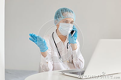 Confused helpless woman doctor wearing gown, surgical mask, medical cap and gloves, posing in front of notebook, at desk, talking Stock Photo