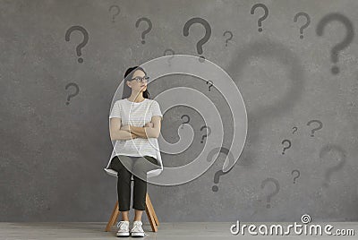 Confused woman sitting on chair and looking at lots of question marks on grey background Stock Photo