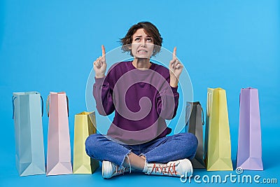 Confused young pretty dark haired curly female with short haircut keeping her index fingers raised while looking upwards with Stock Photo