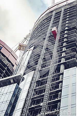 Construction of modern skyscraper building with glass facade. industrial cranes on top and elevator for lifting heavy goods and Stock Photo