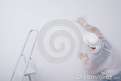 A construction worker in protective clothing and a hard hat, levels the wallpaper with a plastic spatula, to remove excess glue Stock Photo