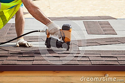 Construction worker putting the asphalt roofing shingles with nail gun on a new frame house. Stock Photo