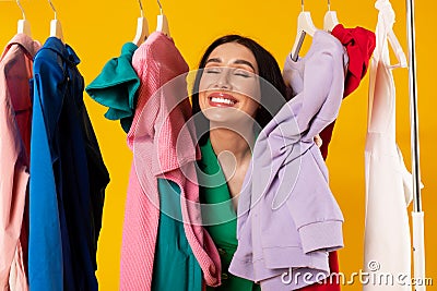 Contented shopaholic woman peeking out between clothes hanging in rail and smiling with closed eyes, yellow background Stock Photo