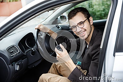 Contented young driver is sitting at his car while looking at the camera. He is holding the keys at his left hand. His right hand Stock Photo