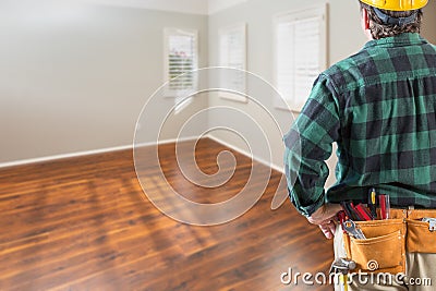 Contractor Wearing Toolbelt and Hard Hat Facing Empty Room with Hard Wood Floors Stock Photo