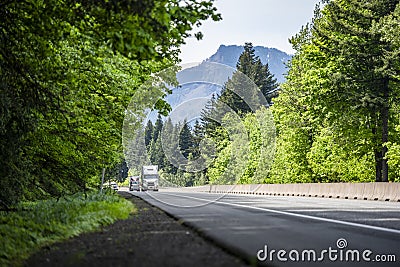 Convoy of different loaded big rigs semi trucks with semi trailers climbing uphill on the forested highway road Stock Photo