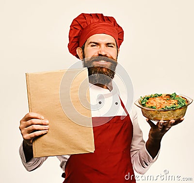 Cook with proud smile in burgundy uniform holds baked dish and menu. Man with beard isolated on white background. Chef Stock Photo