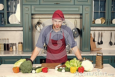 Cook stand at kitchen table. Man in chef hat and apron in kitchen. Vegetables and tools ready for cooking dishes Stock Photo