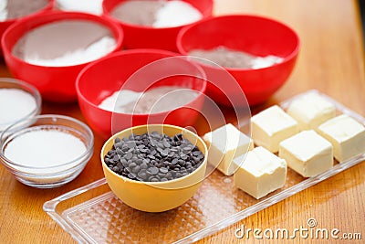 Cookery set of colorful bowls with chocolate drops, flour, cocoa powder, sugar and blocks of butter stand at light brown wooden ta Stock Photo