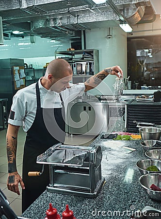 Cooking process. Side view of handsome young chef with black tattoos on his arms pouring flour on kitchen table. Stock Photo