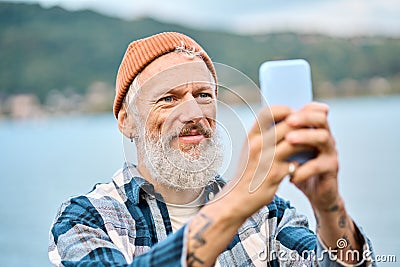 Happy older hipster man standing in nature park taking photos on smartphone. Stock Photo