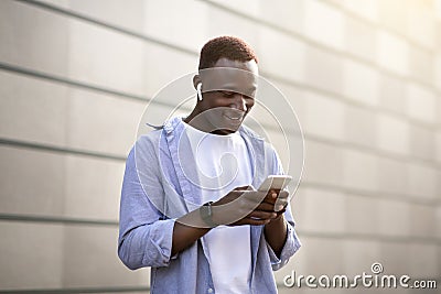 Cool black guy in earphones picking music playlist on mobile phone near brick wall on city street Stock Photo