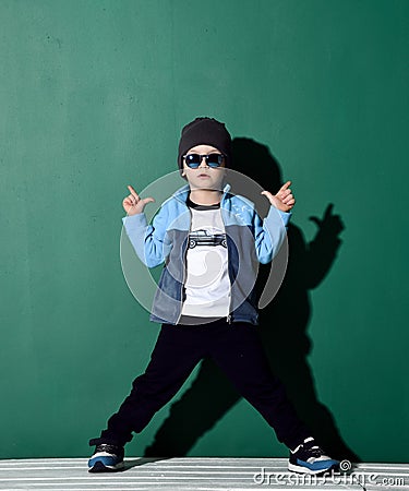 Cool boy in blue sunglasses, headwear, fleece jacket, pants and sneakers is posing like a gangster with two finger guns Stock Photo