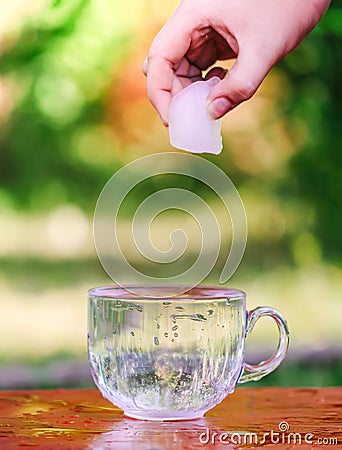 Cool fresh water and ice cube in hand above the transparent glass cup in the table outoors in summer day Stock Photo