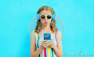 cool girl blowing red lips holding phone listening to music in wireless headphones on colorful blue Stock Photo